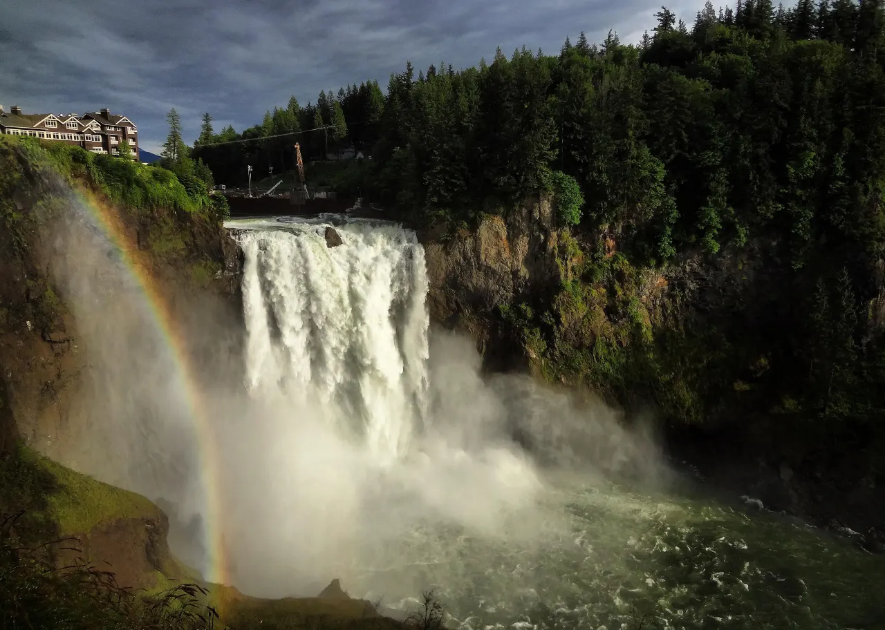 A rainbow in the spray of Snoqualmie Falls in early evening light, the lodge above