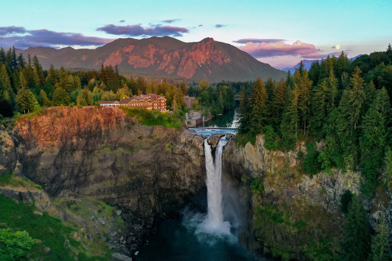 Snoqualmie Falls pouring into its gorge below Salish Lodge, with Mount Si glowing pink at sunset behind