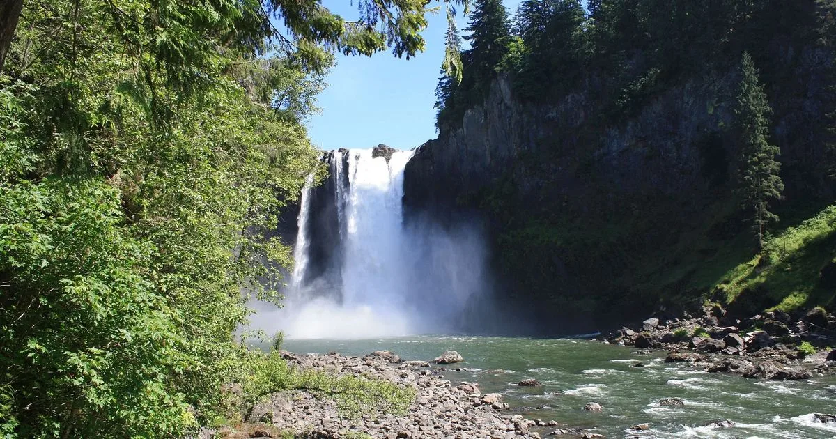 Snoqualmie Falls seen from downriver, framed by green trees, rocky banks and forested cliffs
